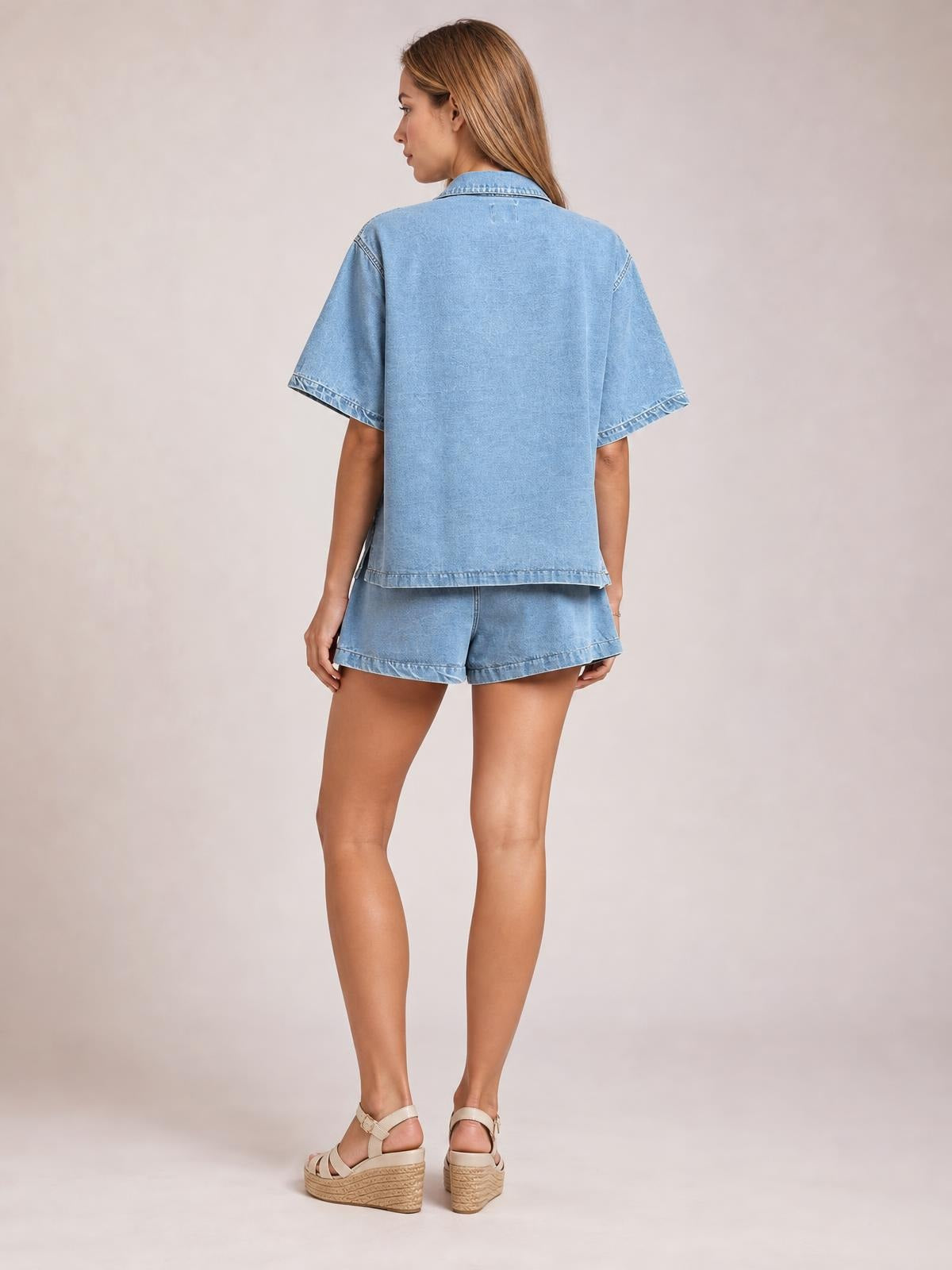 Model viewed from behind wearing a light blue denim button-up top with collared neckline and matching relaxed shorts, standing in a casual pose.
