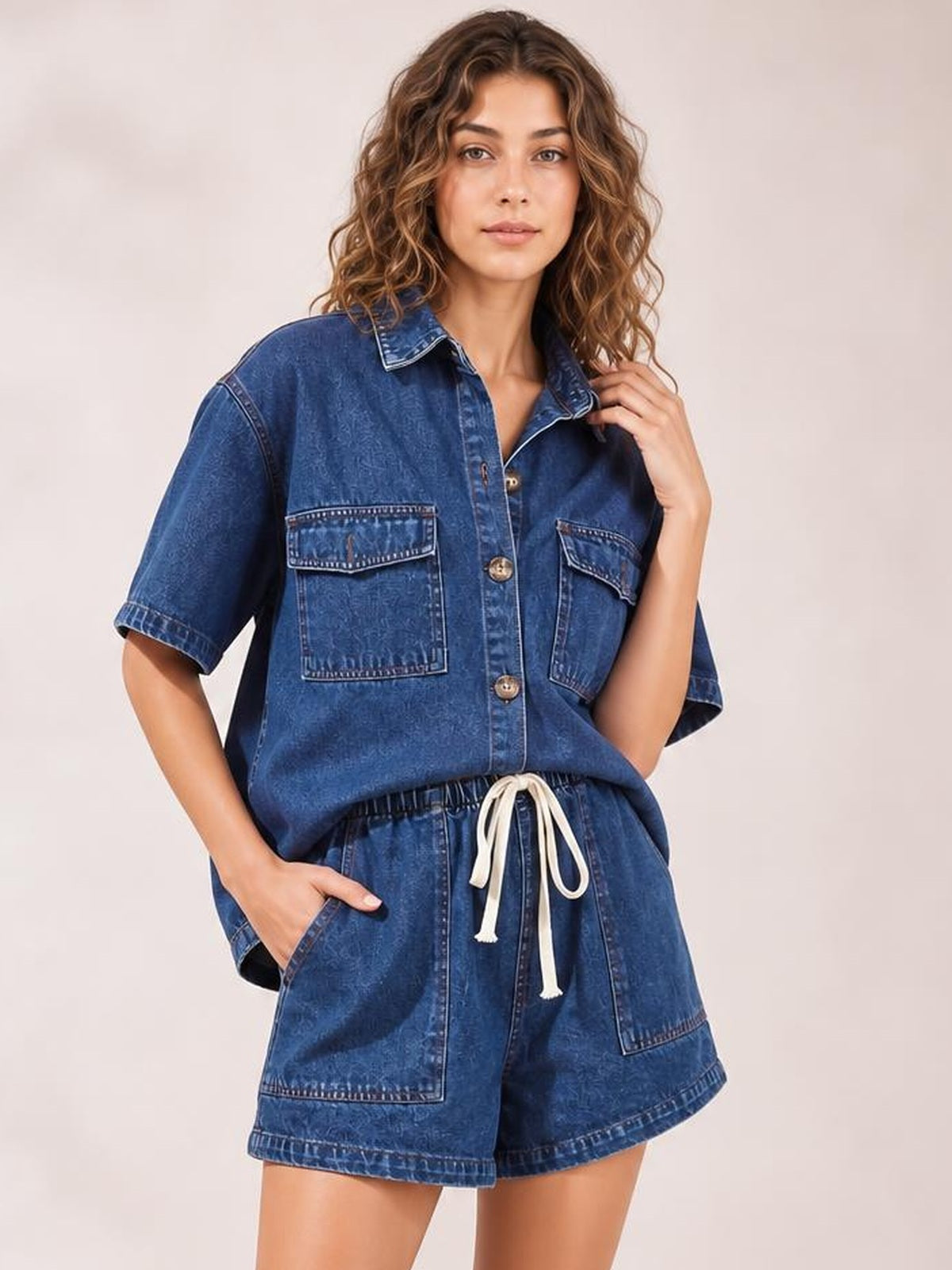 Model poses in a blue denim set with a collared button-up top, flap chest pockets, short sleeves, and matching high-waisted shorts with large pockets.