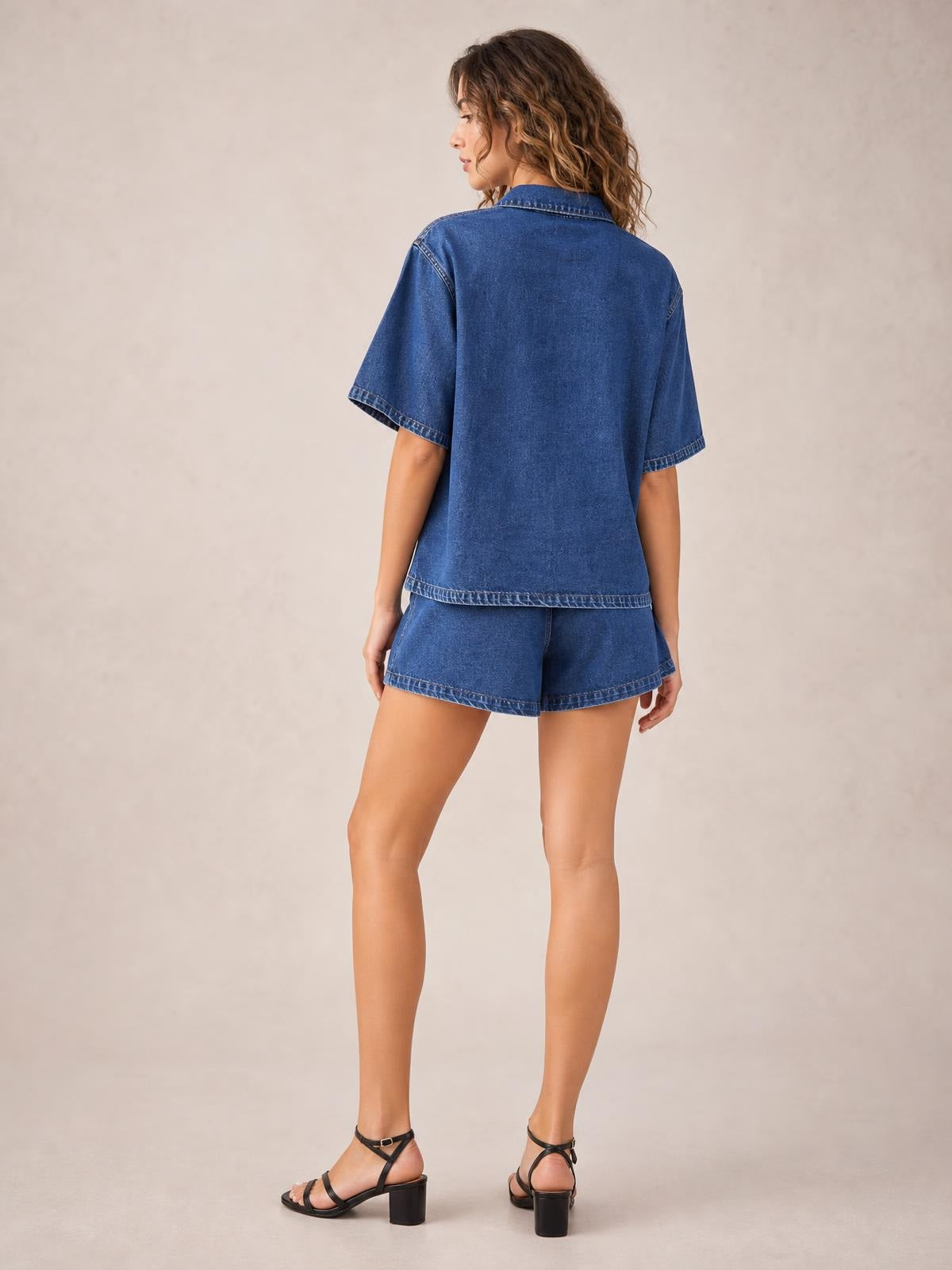 Model shown from behind in a blue denim set with a collared short-sleeve button-up top, relaxed silhouette, matching shorts, and visible contrast stitching.