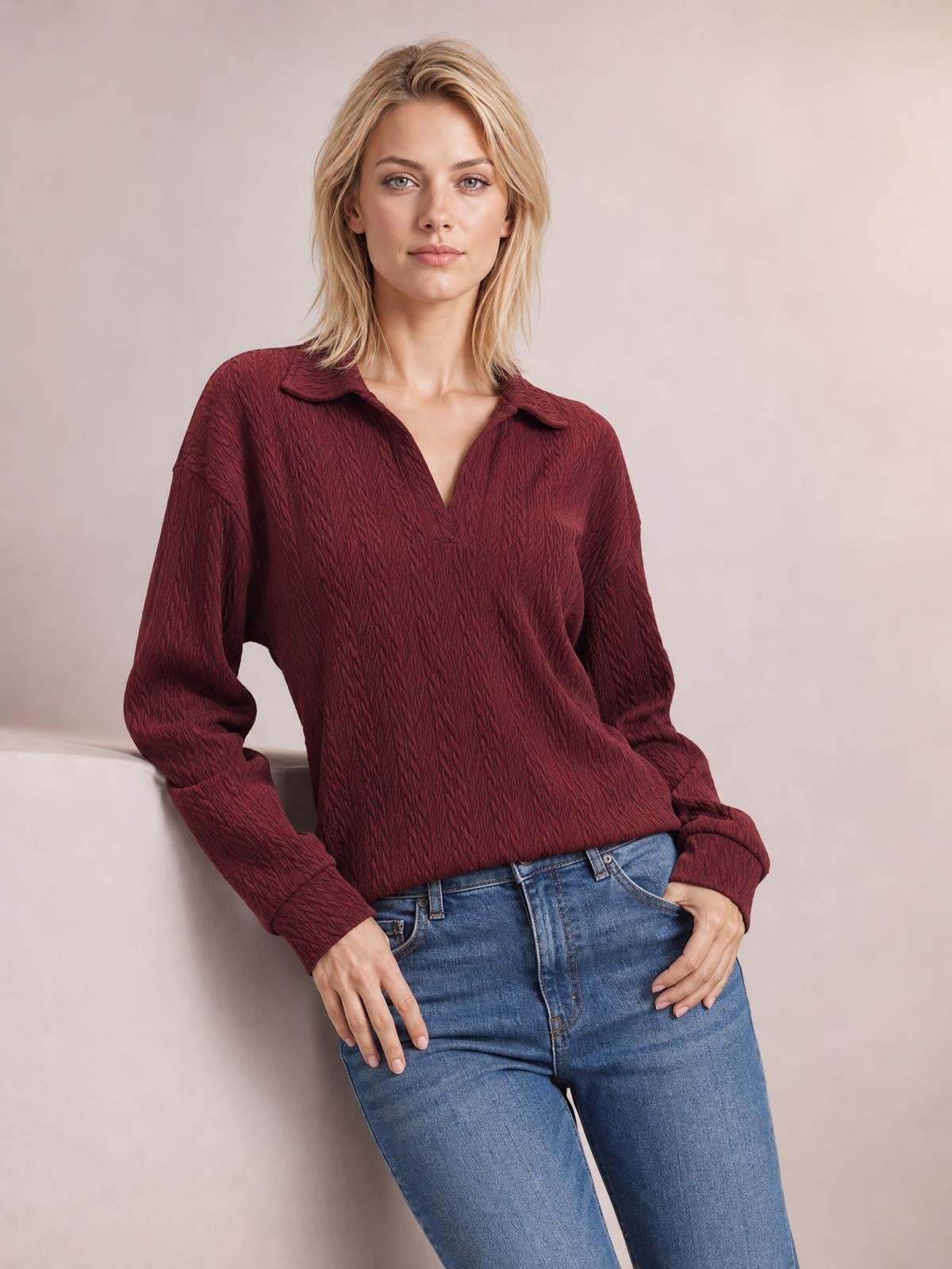 A model wears a solid burgundy cable-knit blouse with a Johnny collar, long sleeves, relaxed regular silhouette, and textured construction while leaning casually.