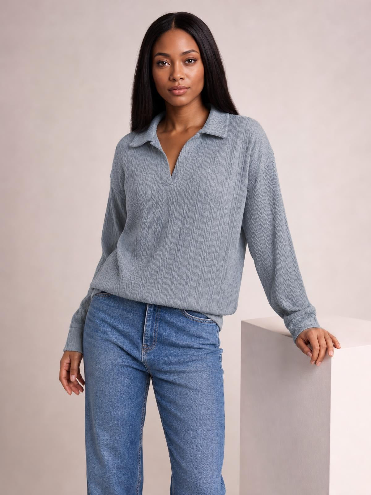 Model wears a blue-gray cable-knit blouse with a Johnny collar, long regular sleeves, and a relaxed regular-length silhouette while leaning on a pedestal.