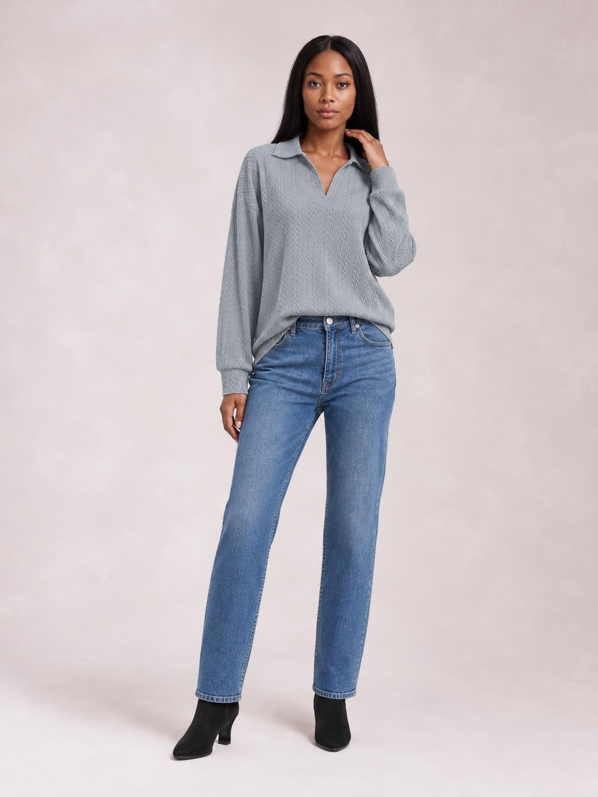 Model wearing a solid gray cable-knit blouse with a Johnny collar, long sleeves, regular silhouette, and curved hem, posed with one hand near her neck.