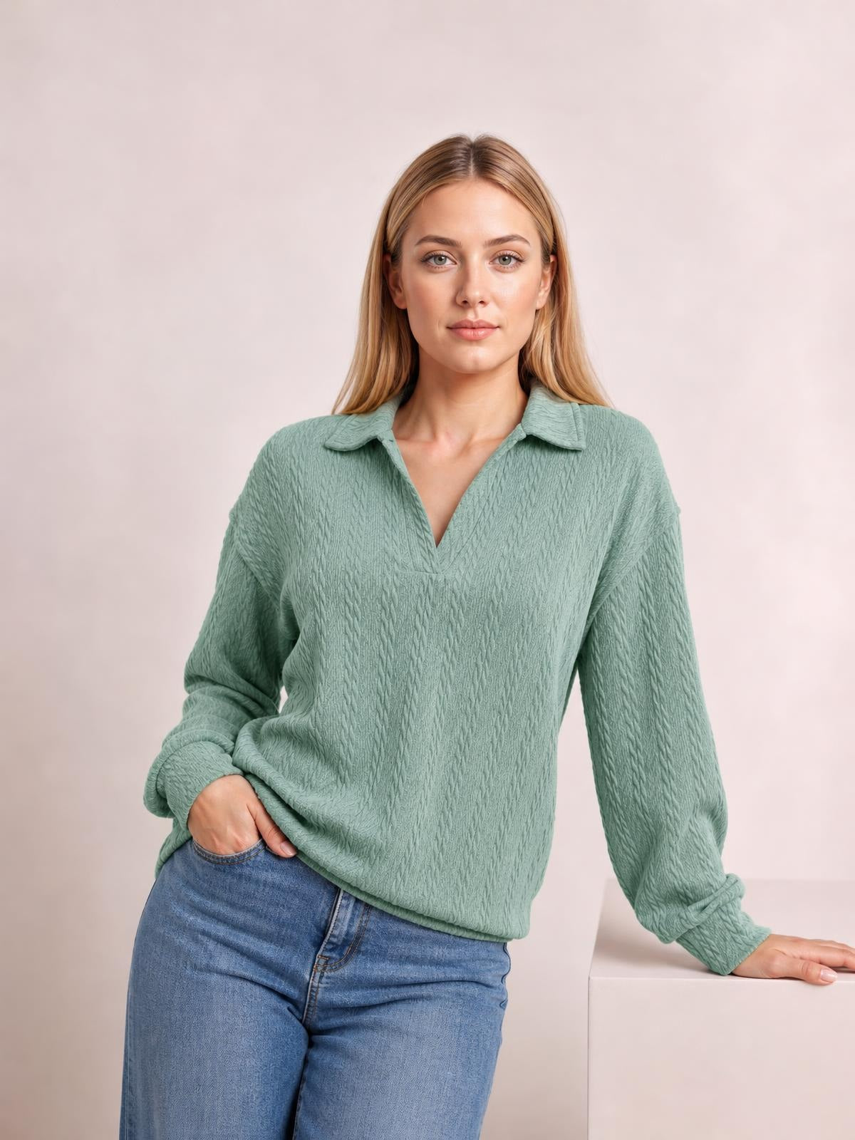 A model wears a sage green cable-knit blouse with a Johnny collar, V-neck opening, regular silhouette, and long sleeves, posing with one hand in her jeans pocket.