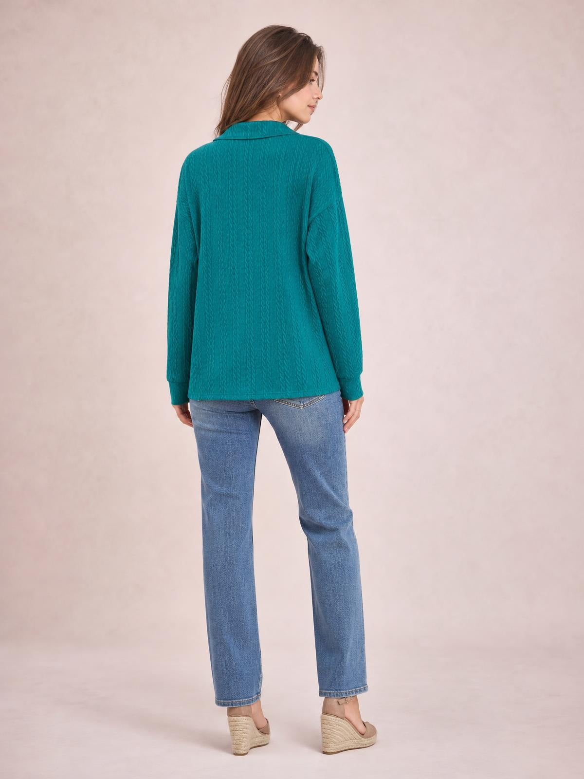 A model looks over her shoulder wearing a teal cable-knit blouse with a collared neckline, long sleeves, and a relaxed regular-length silhouette.