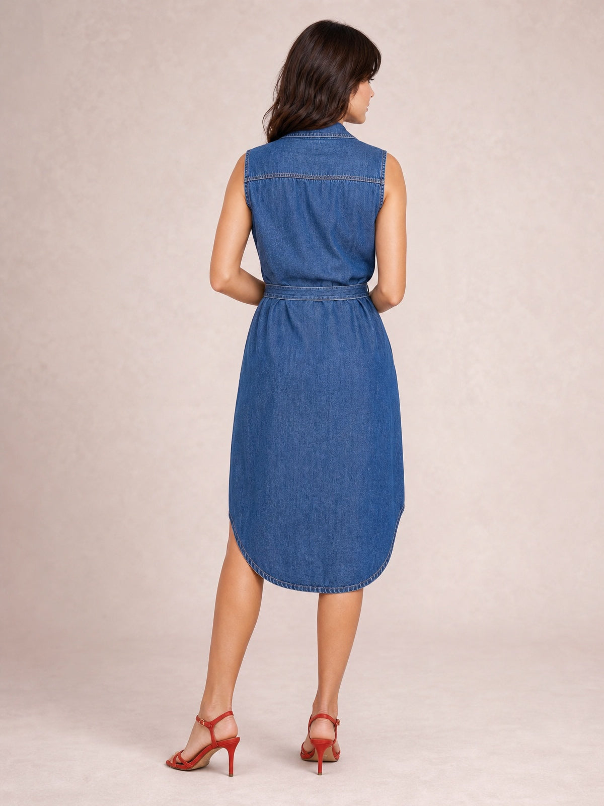 Model shown from behind in a blue collared neck denim dress with cap sleeves, belted waist, visible back seaming, and a softly curved midi-length hem.