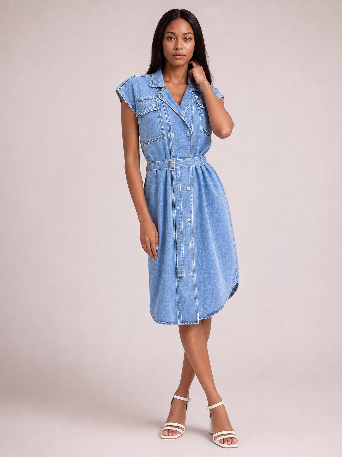 Model poses in a light blue collared neck cap sleeve denim dress with flap pockets, button front, belted waist, and curved midi-length hem.