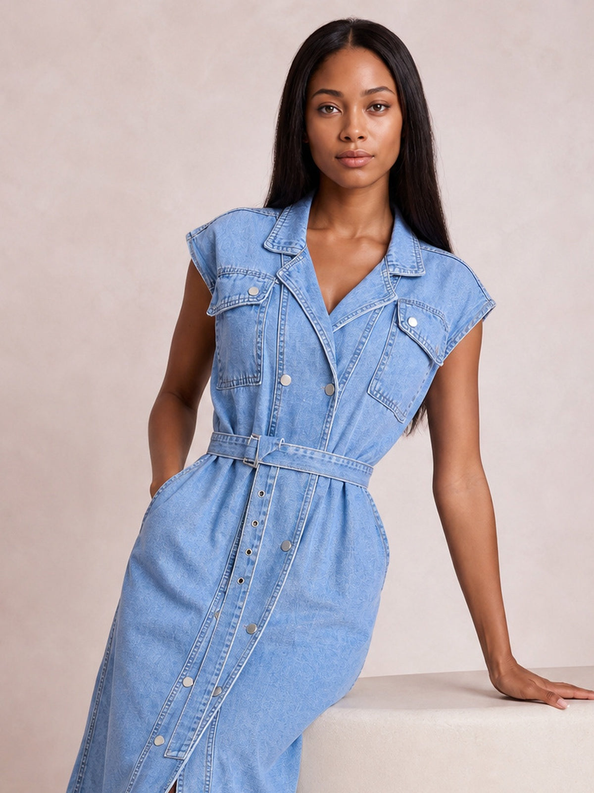 Model leans against a ledge in a light blue collared neck denim dress with cap sleeves, chest pockets, button front, visible seaming, and a belted waist.