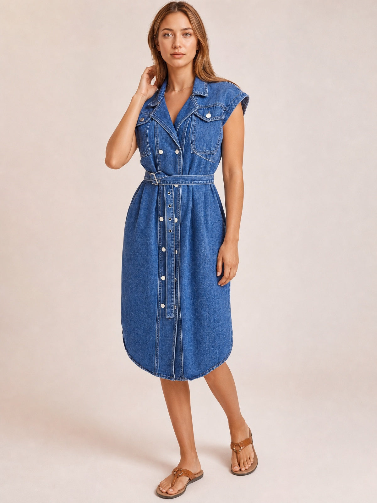 A model poses in a blue collared cap-sleeve denim dress with a belted waist, chest pockets, button-front closure, seamed construction, and a softly curved midi hem.