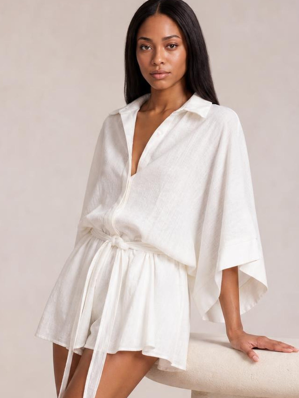 A model leans on a seat wearing a white collared neck half-sleeve romper with a button-down front, tie waist, and relaxed silhouette.