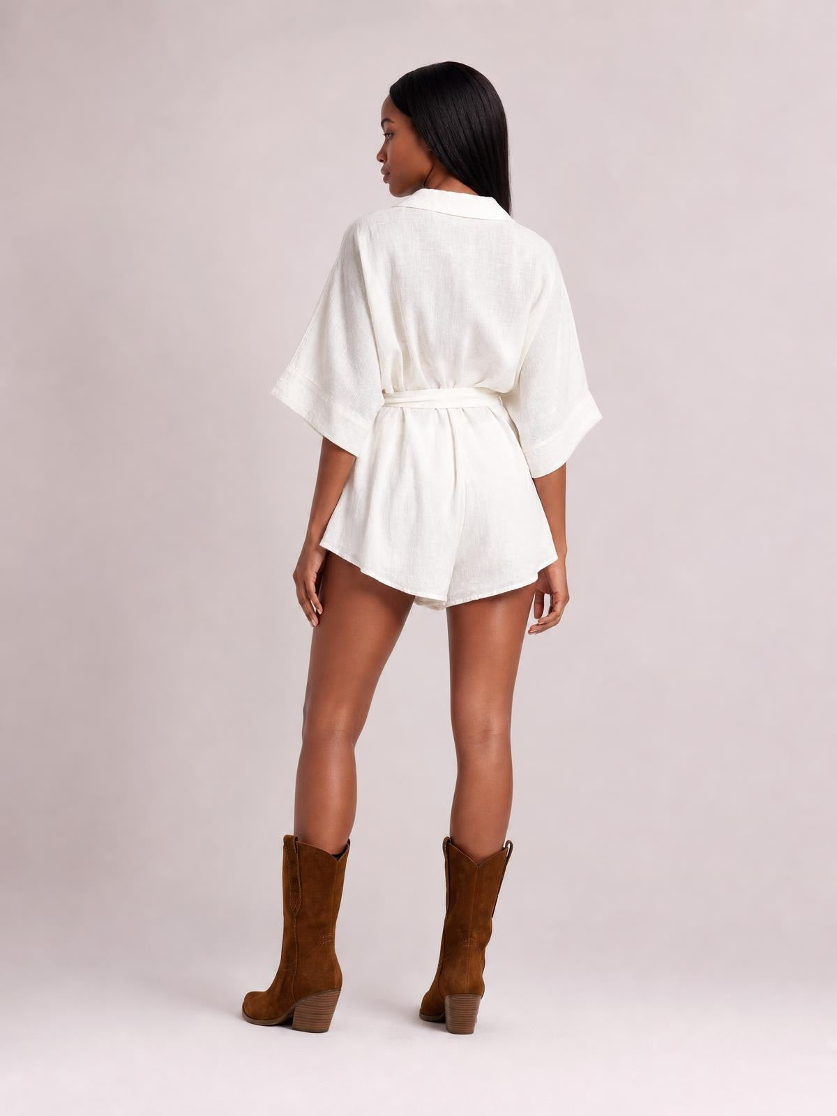 A model stands turned away in a white collared neck half sleeve romper with a tied waist, relaxed silhouette, and short flared legs.