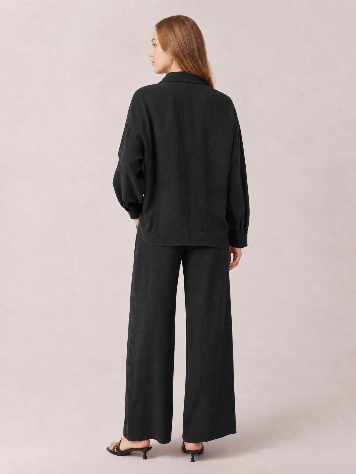 A model stands in a back-facing pose wearing a black collared long-sleeve top and matching wide-leg pants with a relaxed, streamlined silhouette.