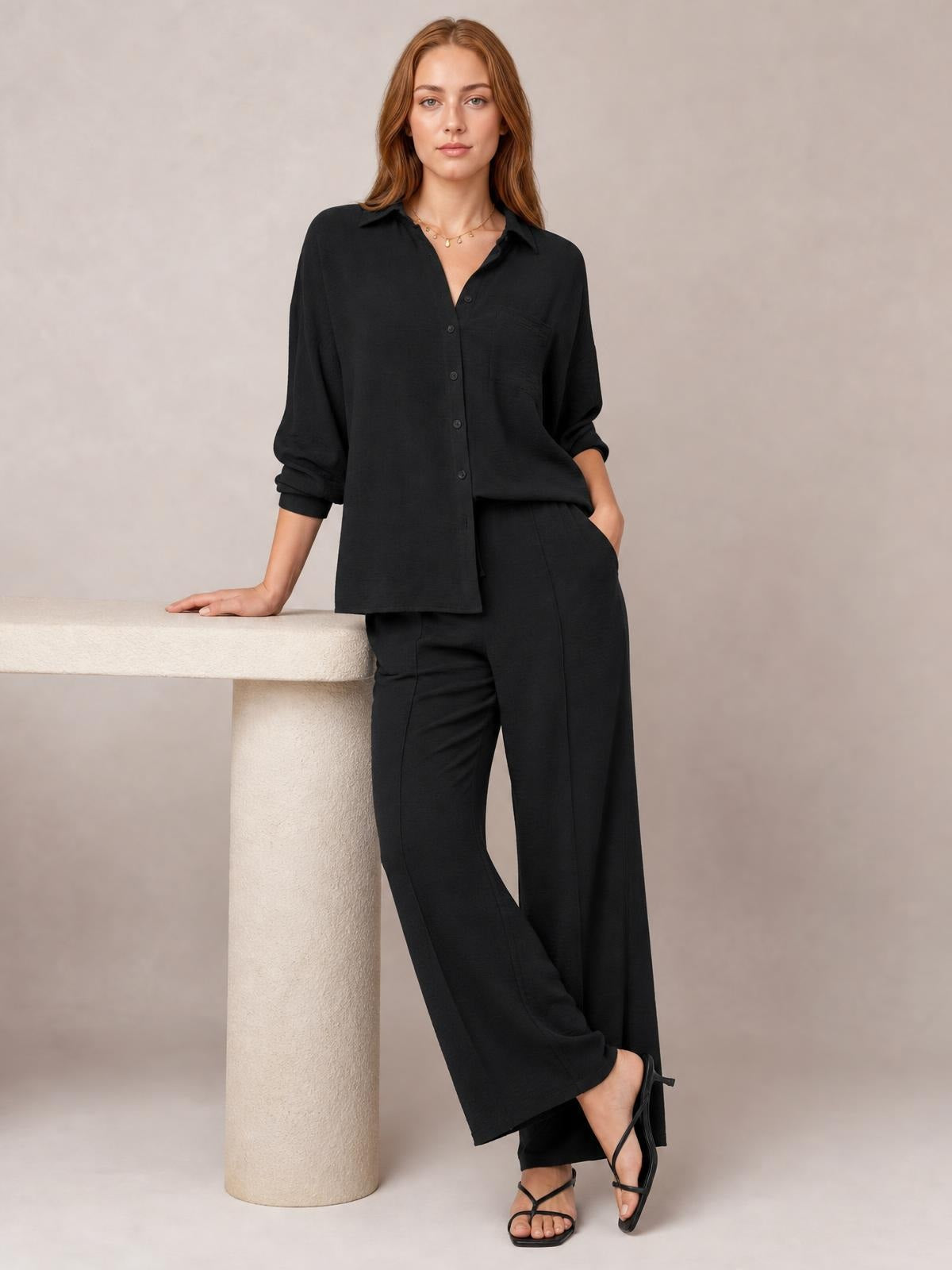 A model poses in a black collared long-sleeve top with button front and matching wide-leg pants, one hand in a pocket and the other resting on a table.