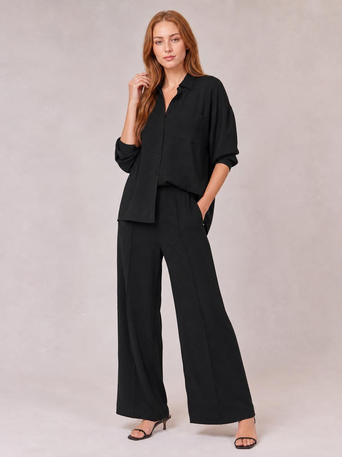 A model poses in a black collared long-sleeve button top and matching pocketed wide-leg pants, with one hand in a pocket.