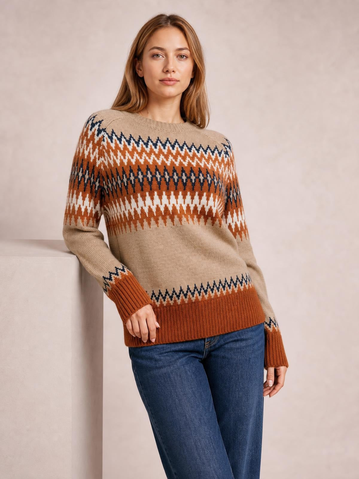 Model leans against a ledge wearing a relaxed crew neck sweater with beige, rust, navy, and white color-blocked geometric patterns, ribbed cuffs and hem.