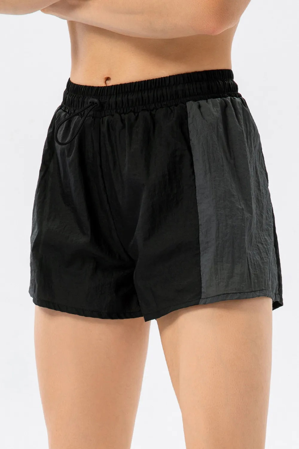 A cropped model wears black color block active shorts with a relaxed silhouette, elastic drawstring waist, and contrasting dark gray side panel.