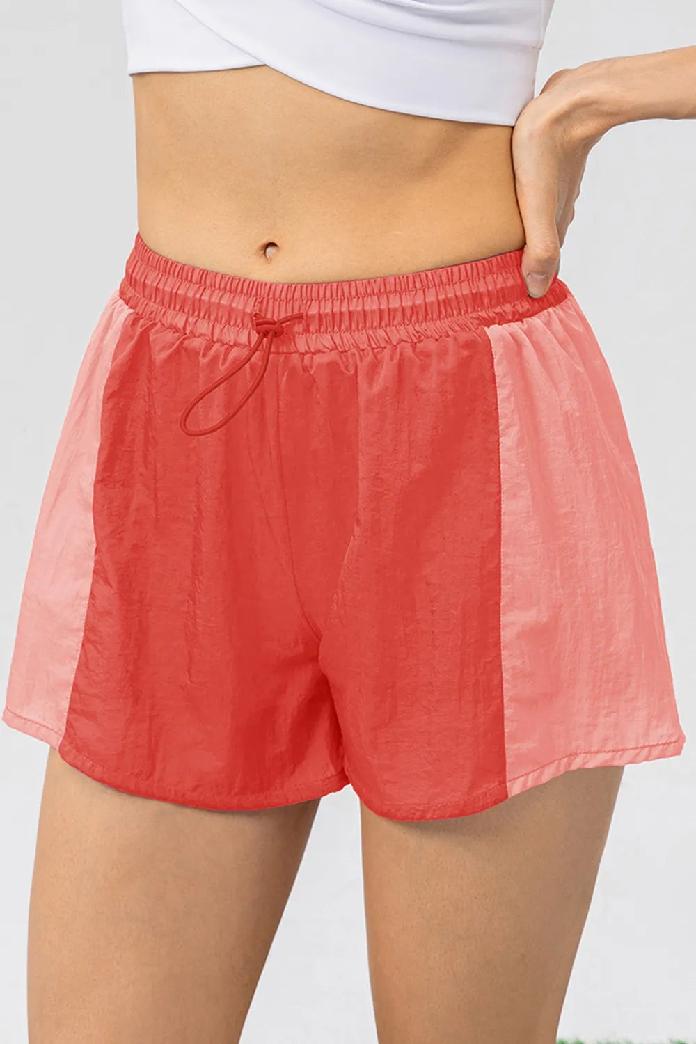 Model wears coral color-block active shorts with a relaxed silhouette, elastic drawstring waist, side panels, and lightweight short hems.