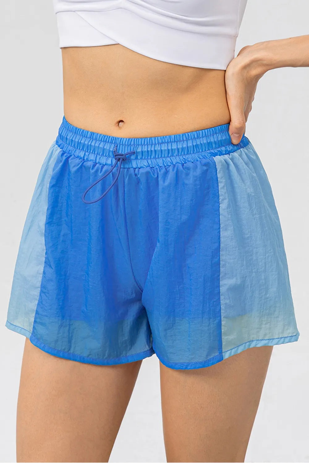 A model wears blue and light blue color block active shorts with a loose silhouette, elastic drawstring waistband, contrast panels, and curved hems.