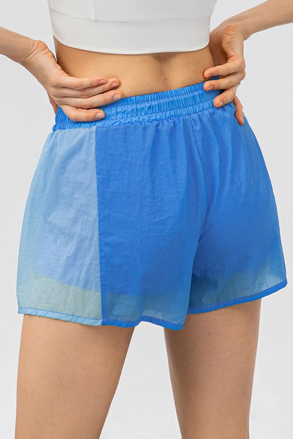 A cropped model adjusts the elastic drawstring waistband of blue color block active shorts with light and bright blue panels and a relaxed short silhouette.