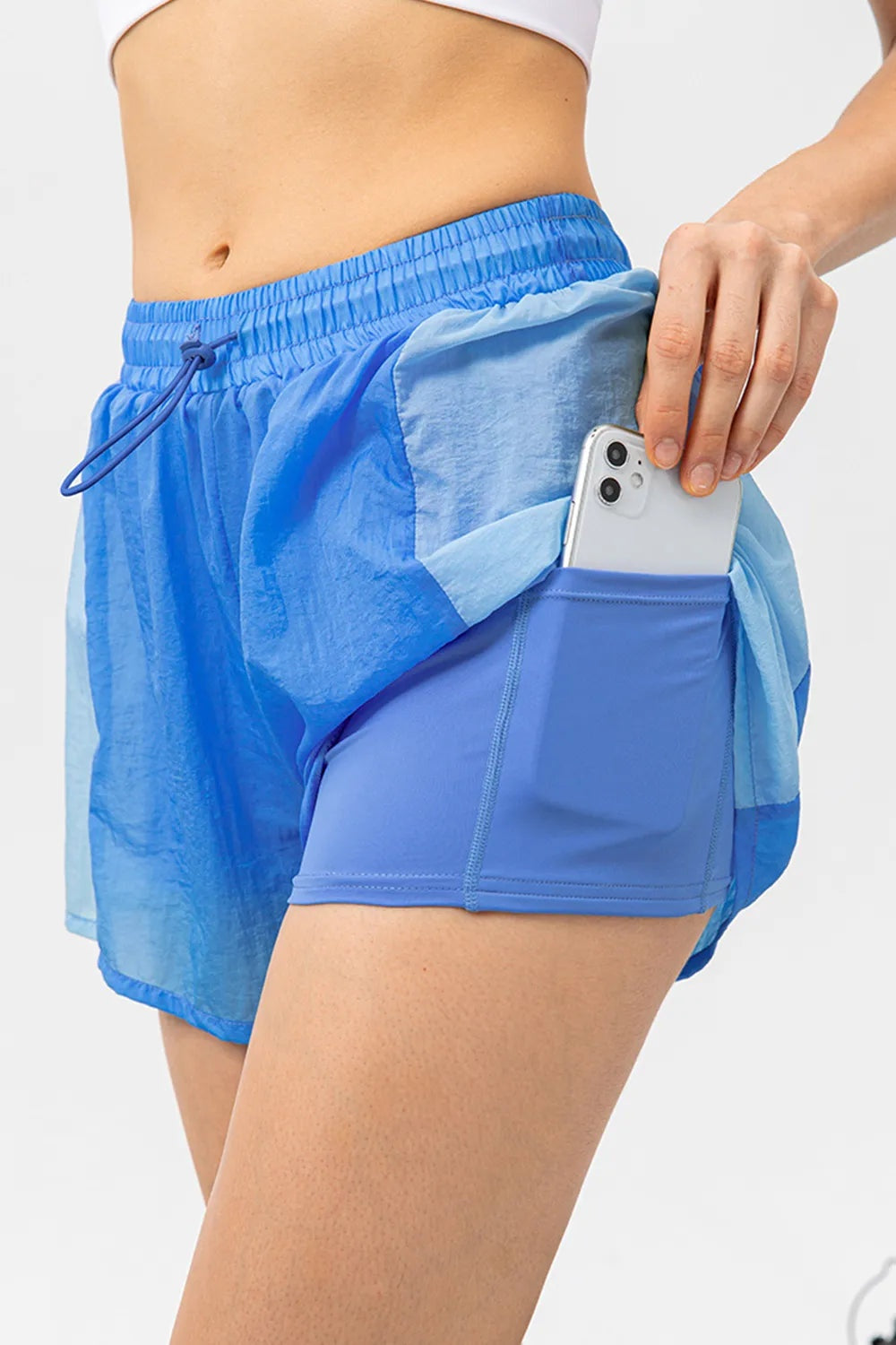 Visible model wears blue color-block active shorts with an elastic drawstring waist, layered panels, and a fitted inner pocket short while holding a phone.