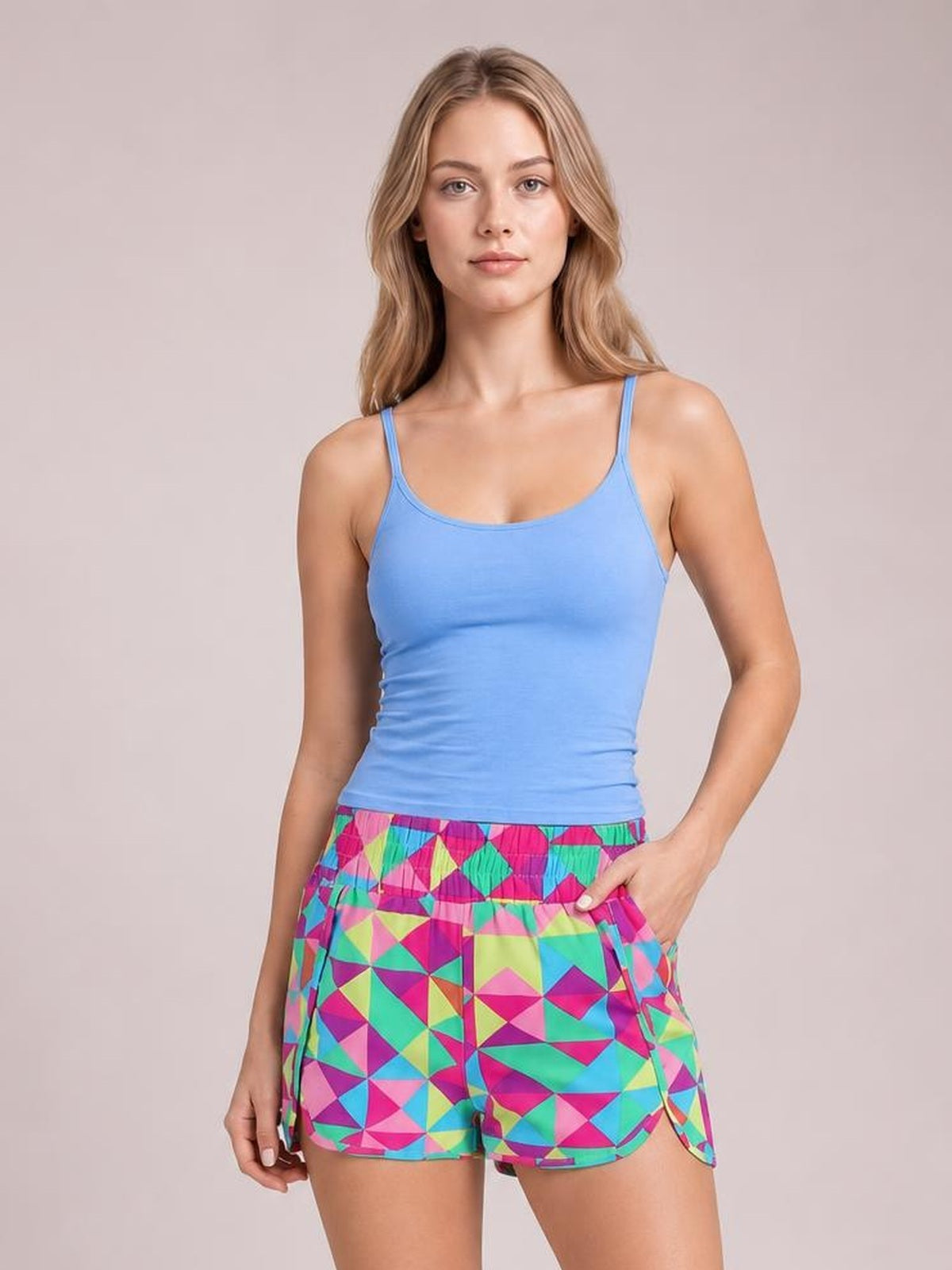 A woman modeling a bright blue ribbed tank top and colorful geometric print athletic shorts against a solid light beige studio background.
