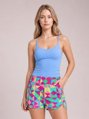A woman modeling a bright blue ribbed tank top and colorful geometric print athletic shorts against a solid light beige studio background.