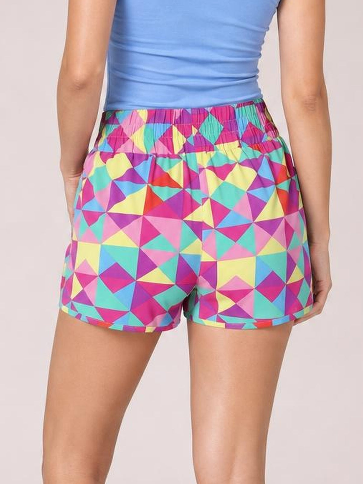 Model shown from behind wearing vibrant geometric color block elastic waist shorts with a relaxed silhouette, curved hems, and opaque polyester construction.
