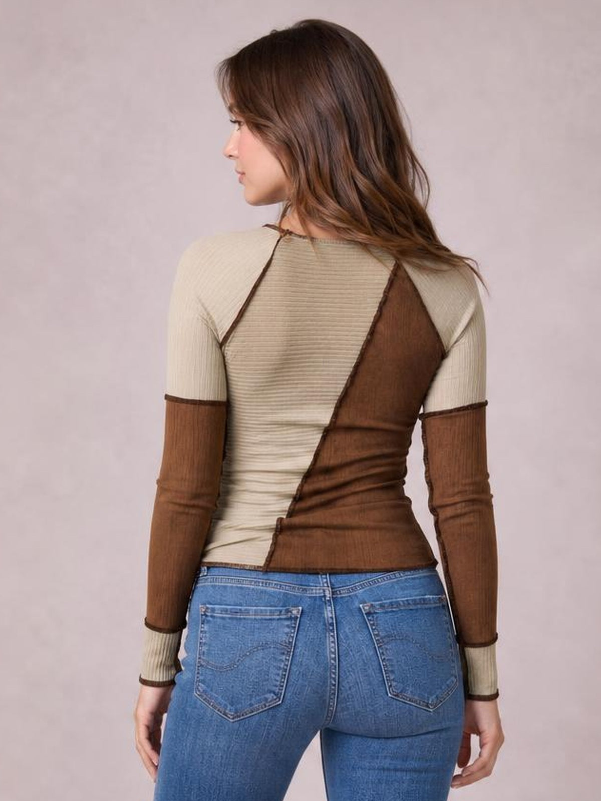 A model wears a fitted color block knit top in cream and brown with ribbed texture, exposed seams, and long raglan sleeves, shown from the back.