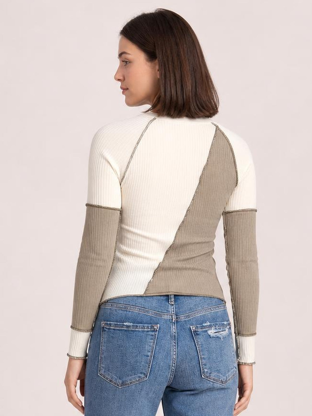 A model shown from behind wears a cream and taupe color block ribbed knit top with exposed seams, long raglan sleeves, and a fitted regular-length silhouette.