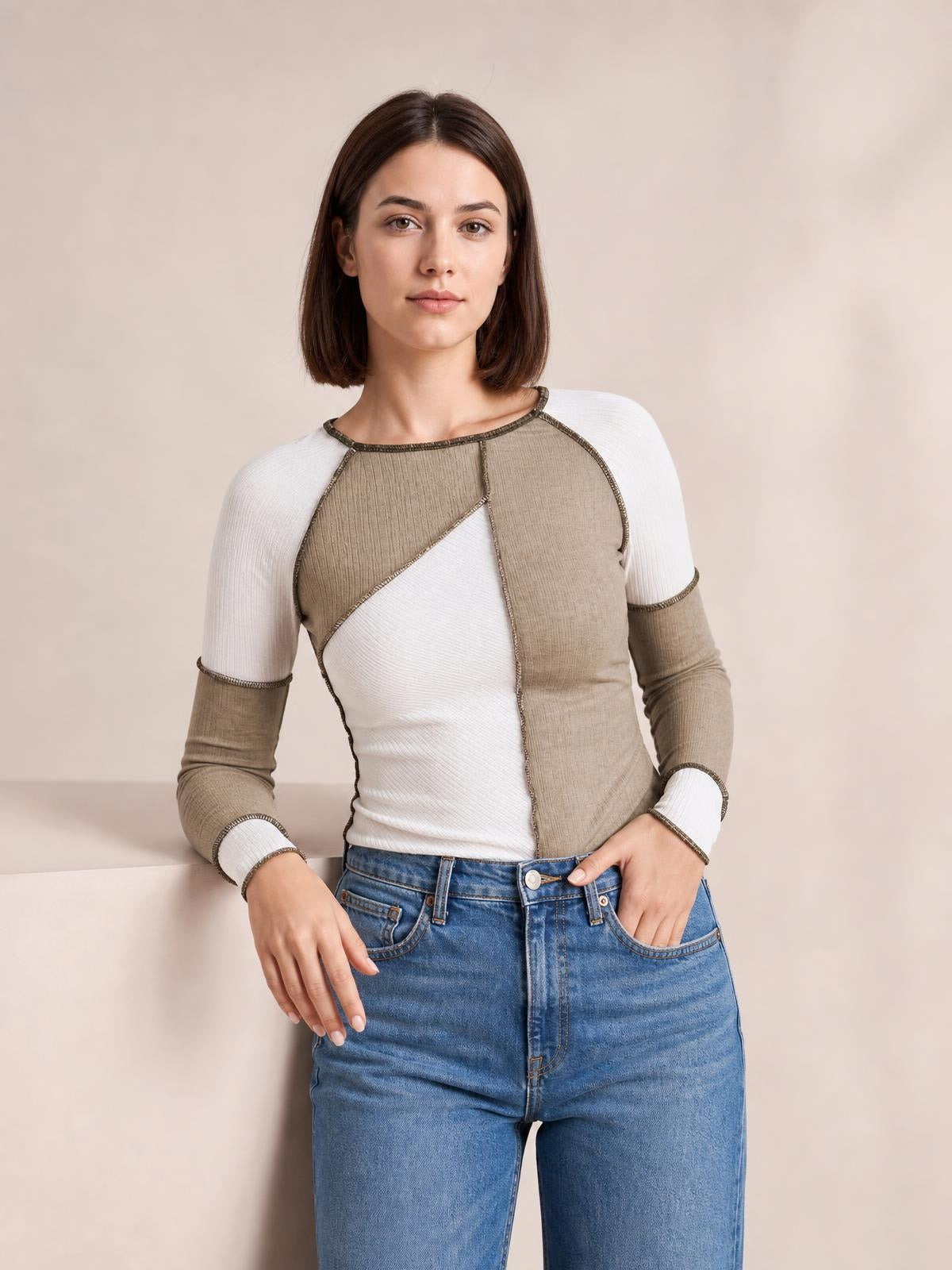 Model wearing a taupe and ivory color block knit top with a round neck, ribbed texture, exposed seams, and long raglan sleeves, posed with one hand in her jeans pocket.