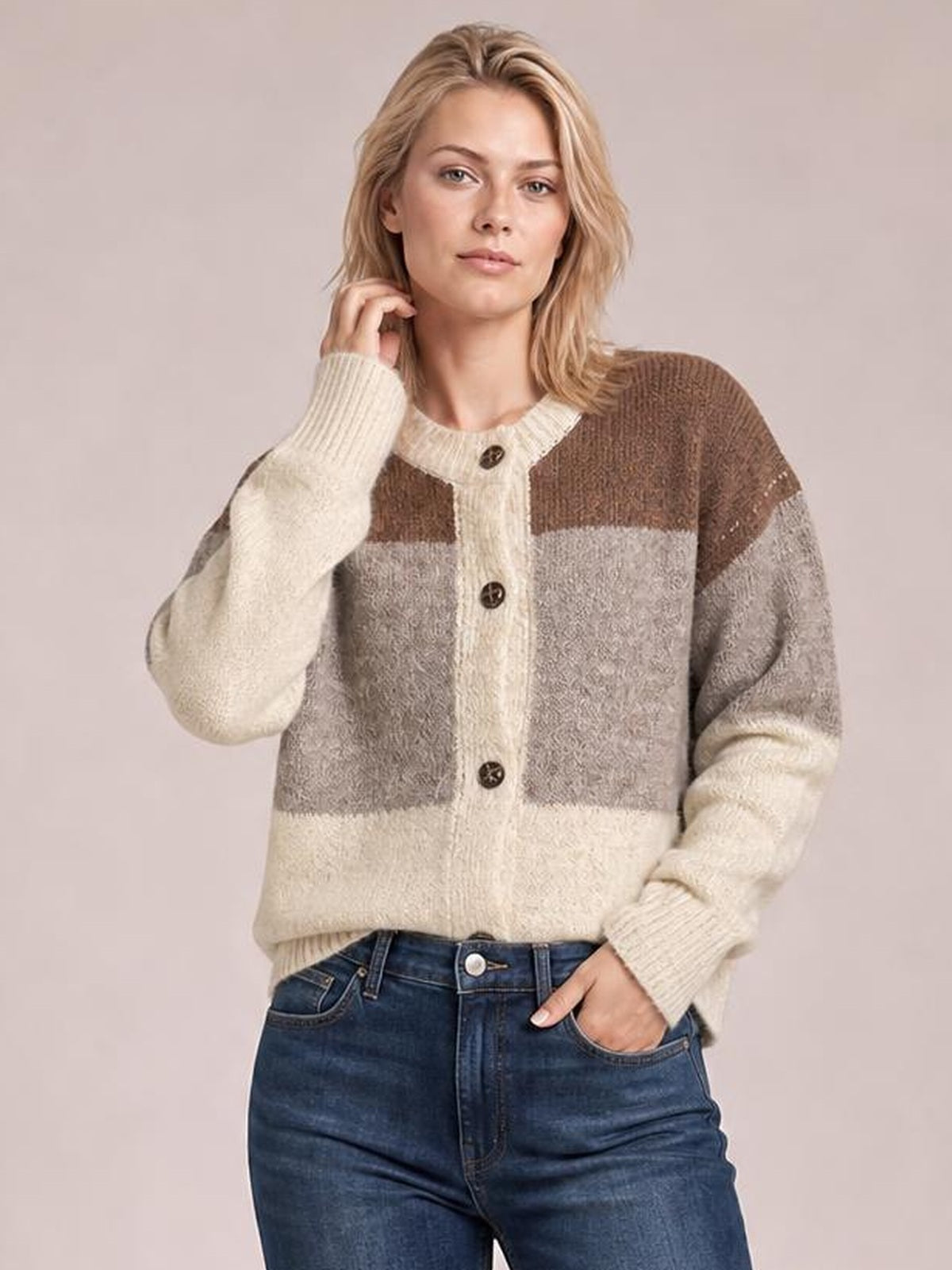 Model poses in blue jeans wearing a relaxed color block round neck button-up cardigan with cream, gray, and brown panels, ribbed cuffs, and front buttons.