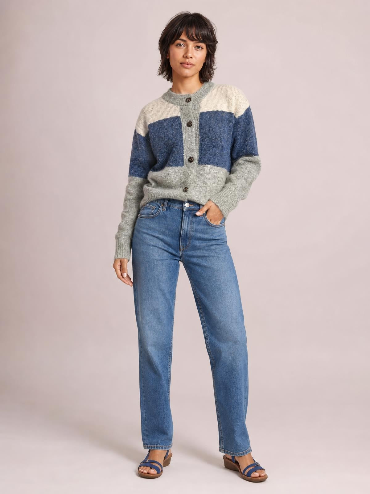 Model poses in a color block round neck cardigan with button-up front, gray, cream, and blue knit panels, long sleeves, and a relaxed silhouette.