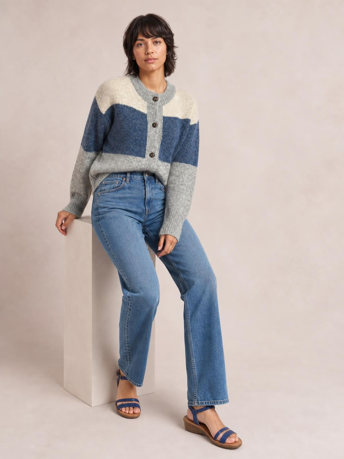 Model sits casually in a relaxed color-block cardigan with a round neck, button front, long sleeves, and cream, blue, and gray sections.