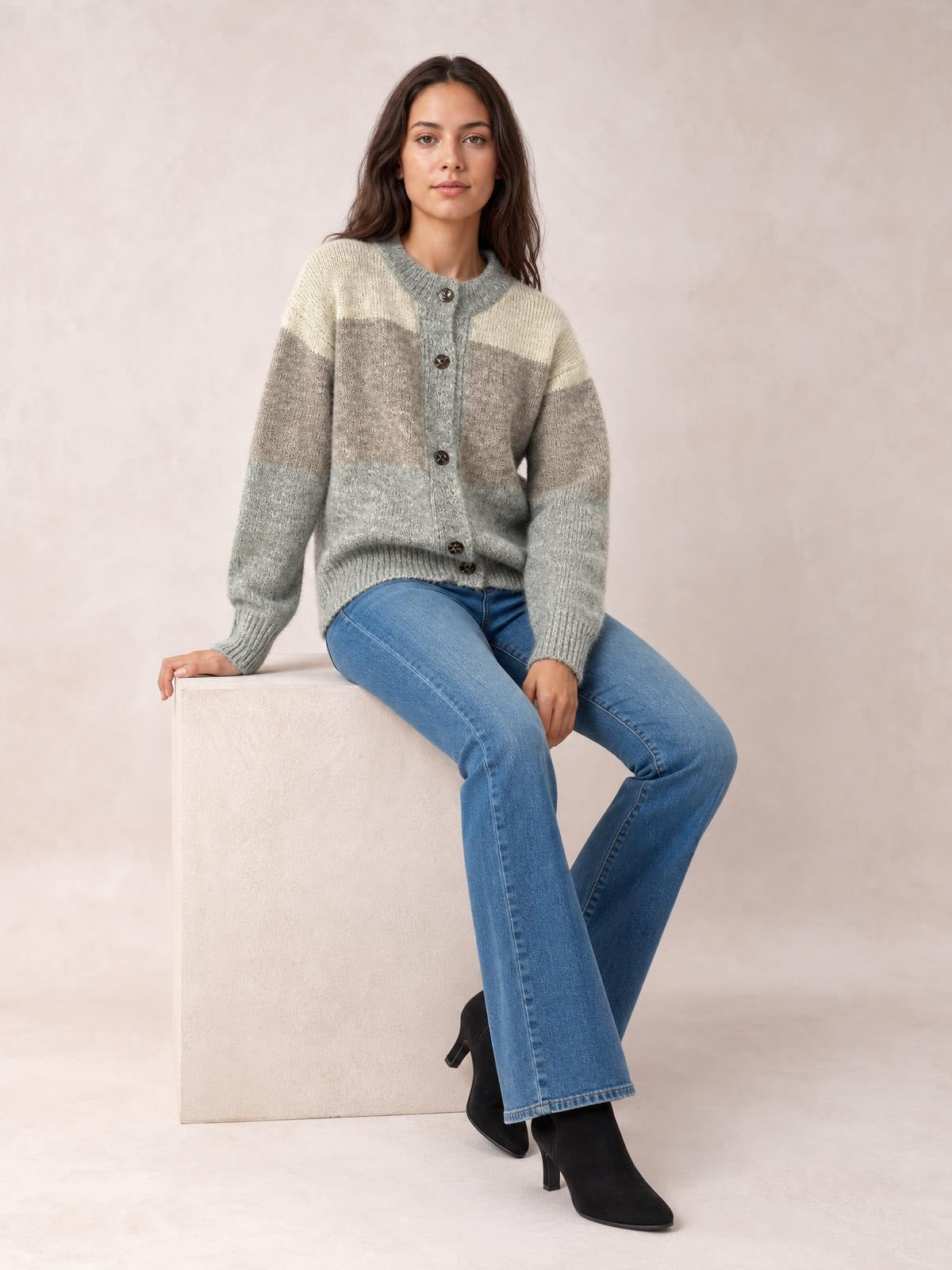 Model seated in blue jeans wears a relaxed color block cardigan with a round neck, button-up front, ribbed trim, and cream, taupe, and gray sections.