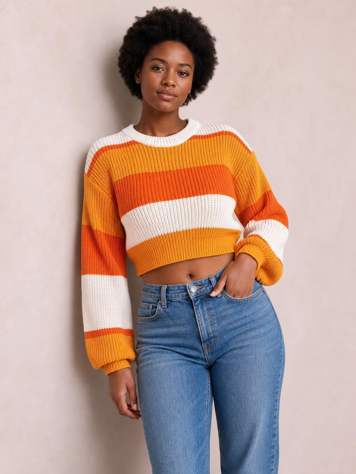 Model wearing a cropped round-neck color block sweater in orange, yellow, and white with ribbed trim, relaxed sleeves, and a hand in her jeans pocket.