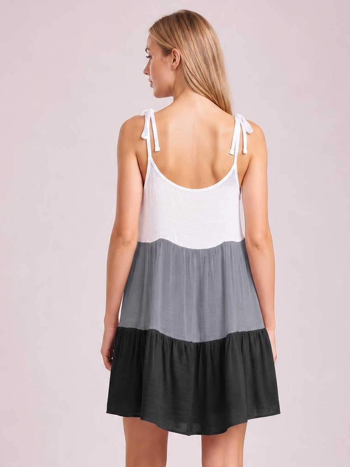 A model faces away in a semi-sheer color block cover-up dress with tied spaghetti straps, a loose tiered silhouette, and white, gray, and black panels.