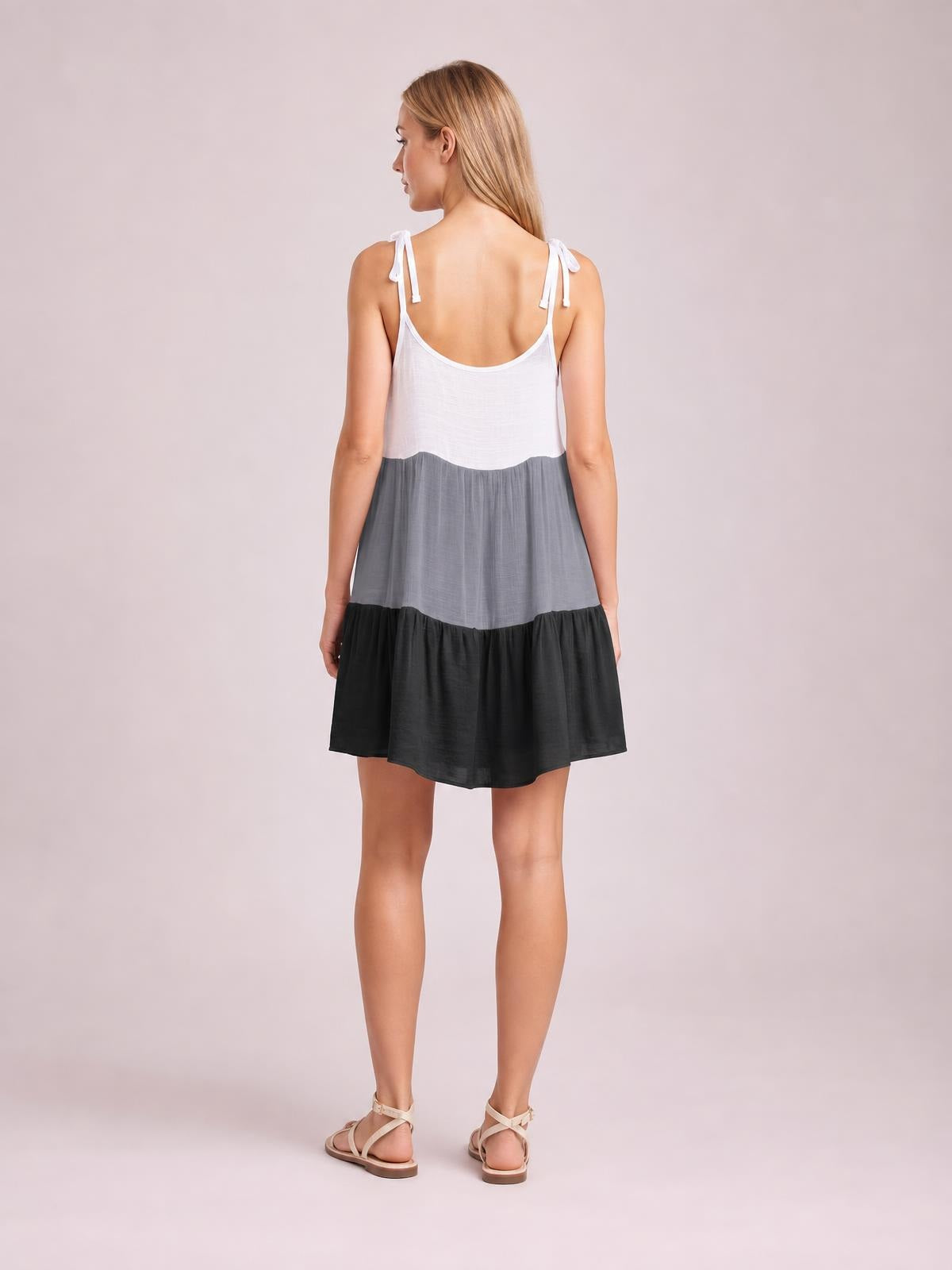 Back view of a model wearing a semi-sheer color block spaghetti strap cover-up dress in white, gray, and black, with tied straps and a loose tiered mini silhouette.
