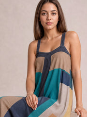 Model seated in a flowing color-block midi dress with a square neckline, wide straps, brown, teal, navy, cream, and golden panels, and a softly flared silhouette.
