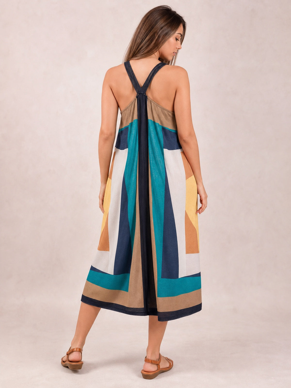 Model shown from the back in a flowing color-block midi dress with wide crossed straps, brown, teal, navy, cream, and golden panels, and a softly flared silhouette.