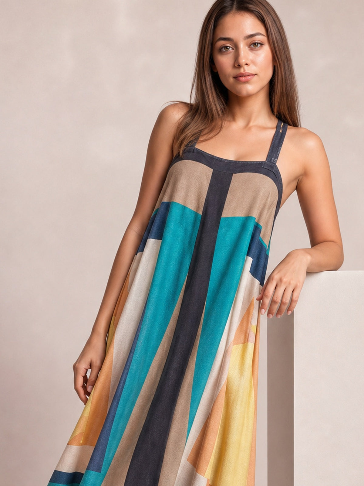 Model poses in a flowing color-block midi dress with a square neckline, wide straps, and bold teal, navy, cream, brown, and golden panels.