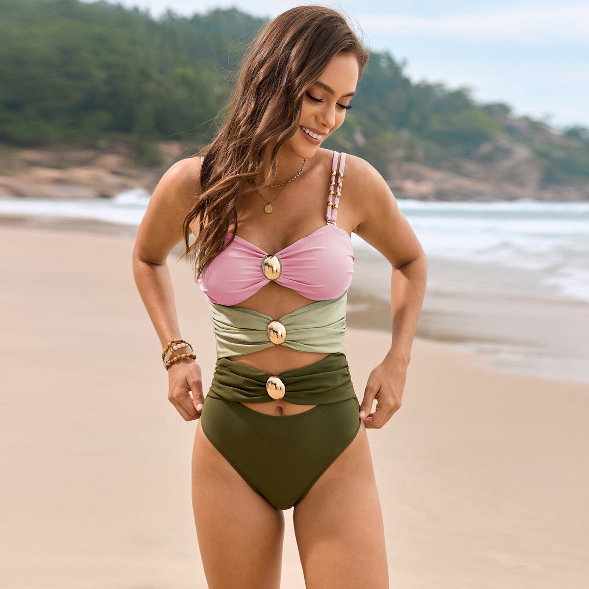 A model stands in a color block one-piece swimsuit with a pink ruched sweetheart-style top, sage and olive panels, cutouts, and gold-tone accents.