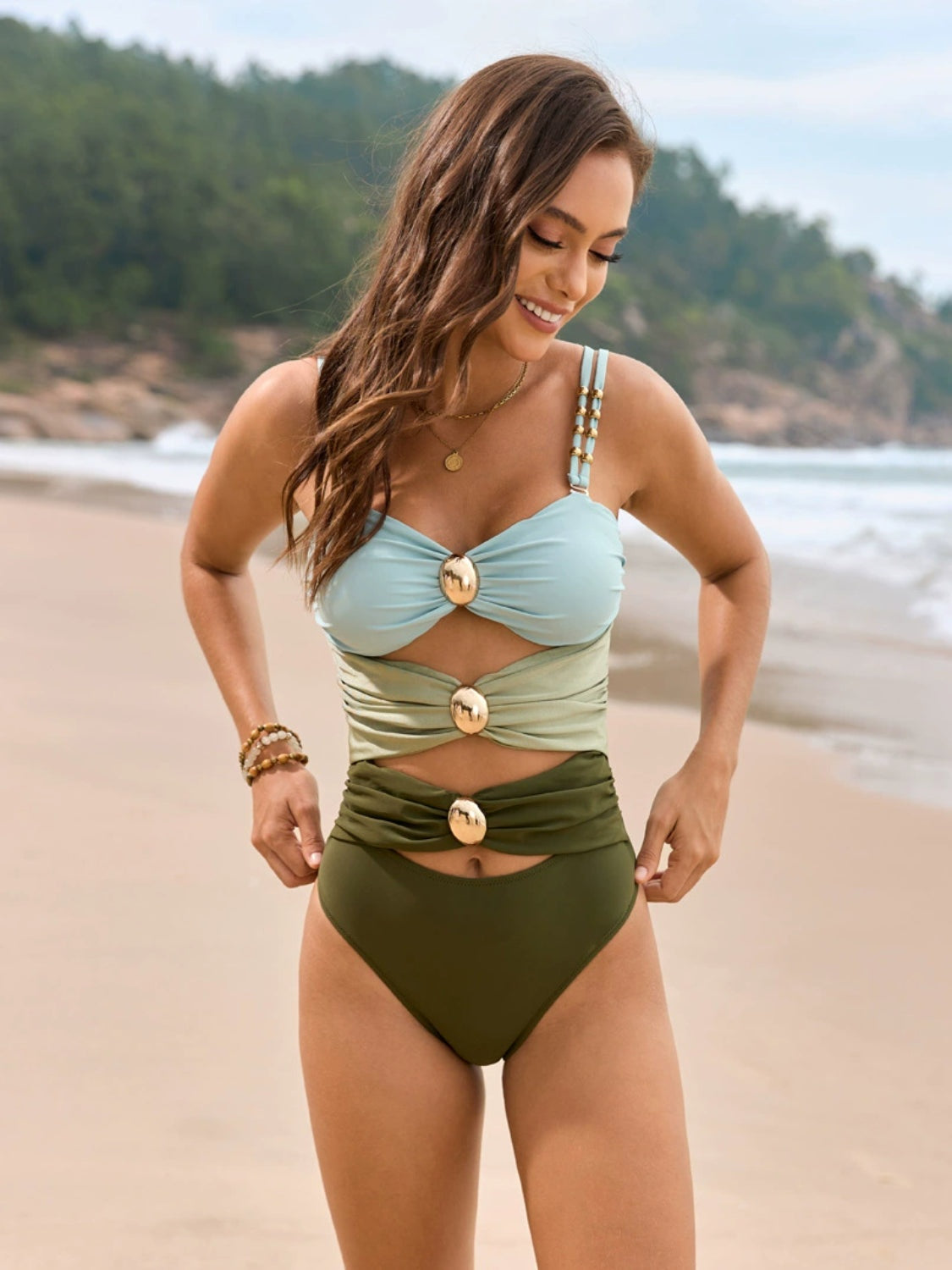 Model poses with hands at her hips in a color block one-piece swimsuit with a ruched sweetheart neckline, gathered panels, gold-tone accents, and a high-waisted silhouette.