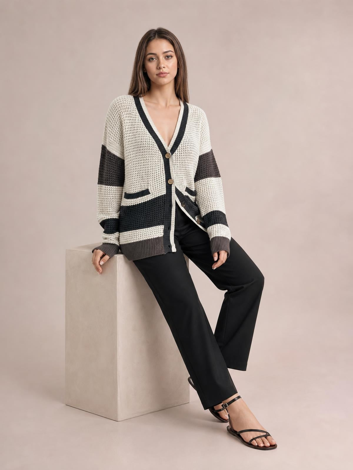 Model seated in a relaxed pose wears a color block V-neck cardigan with waffle-knit texture, button front, contrast trim, striped sleeves, and front pockets.