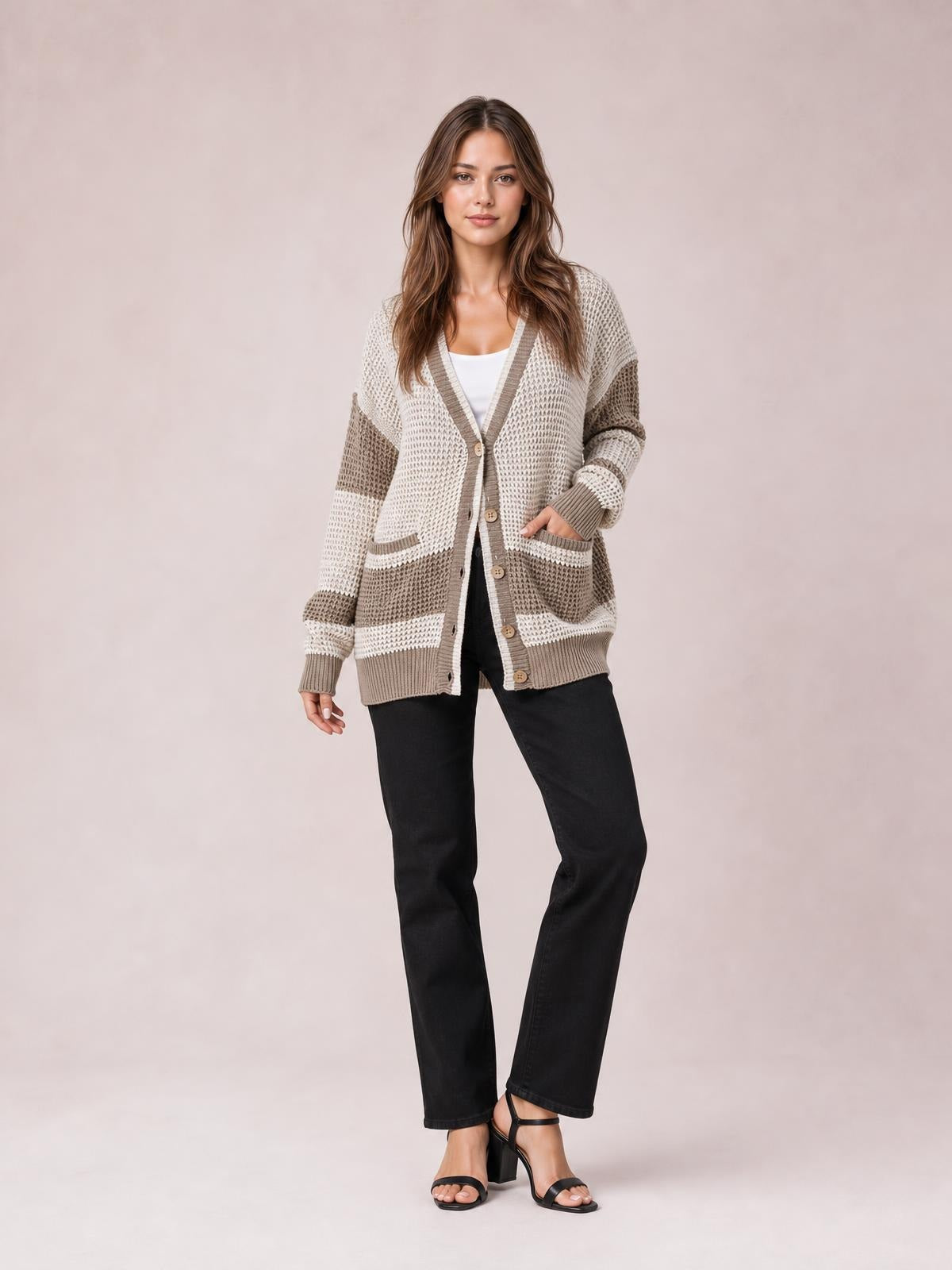 Model stands in a relaxed cream and taupe color block V-neck cardigan with waffle-knit texture, button front, ribbed trim, pockets, and a long silhouette.