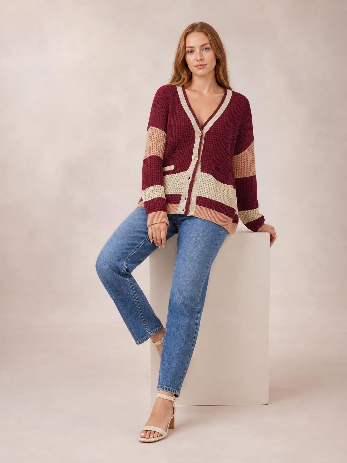 A woman sits in a burgundy and cream color block V-neck cardigan with waffle-knit texture, button front, contrasting bands, pockets, and a relaxed silhouette.