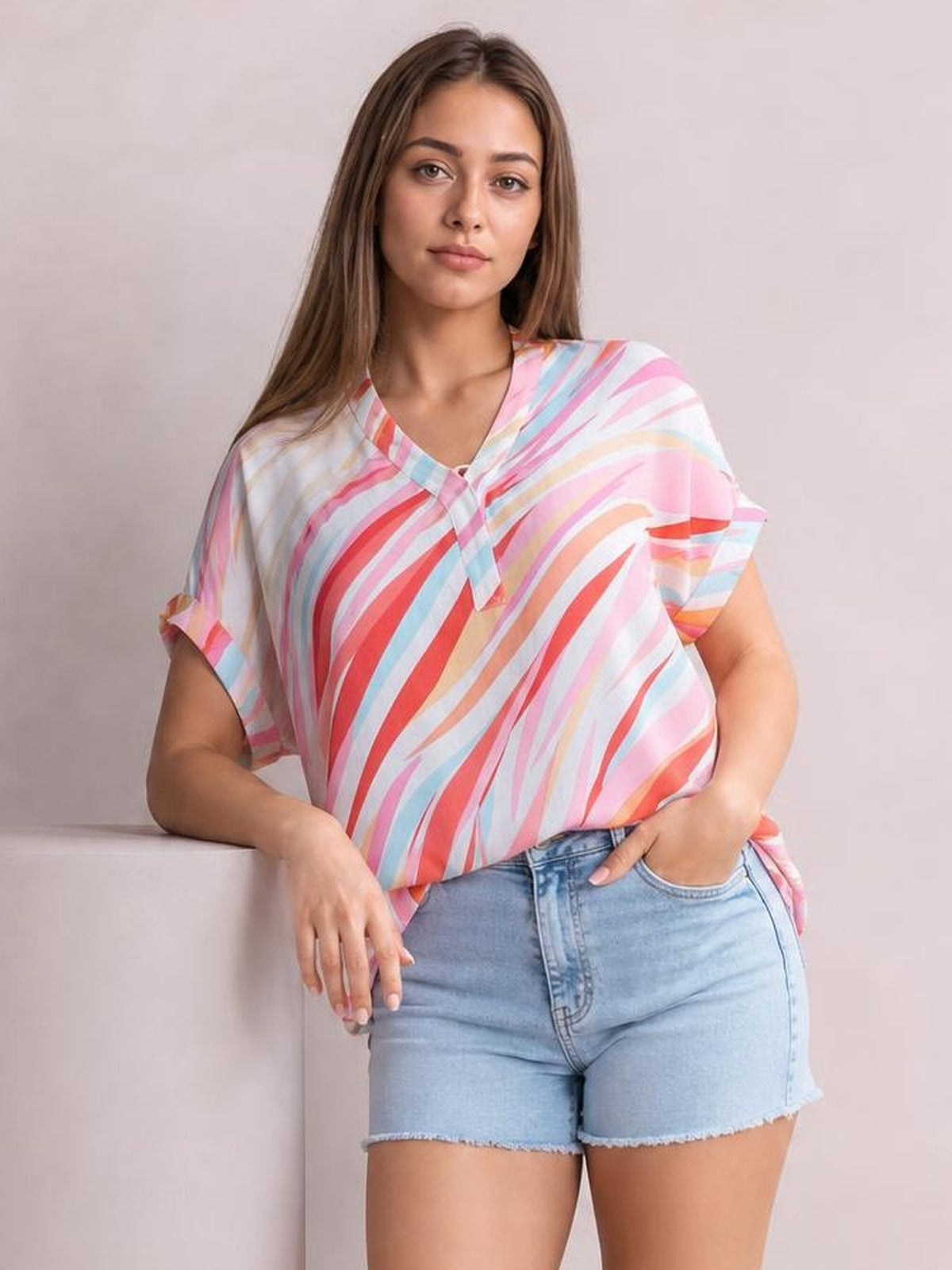 Model wears a colorful striped color-block blouse with a V-neck, loose silhouette, and rolled half sleeves while posing with one hand in her shorts pocket.