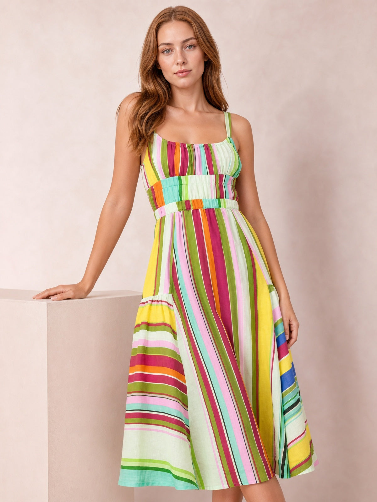 Model poses in a colorful striped spaghetti strap midi dress with a gathered neckline, defined waistband, and softly flared skirt in a bright yellow colorway.