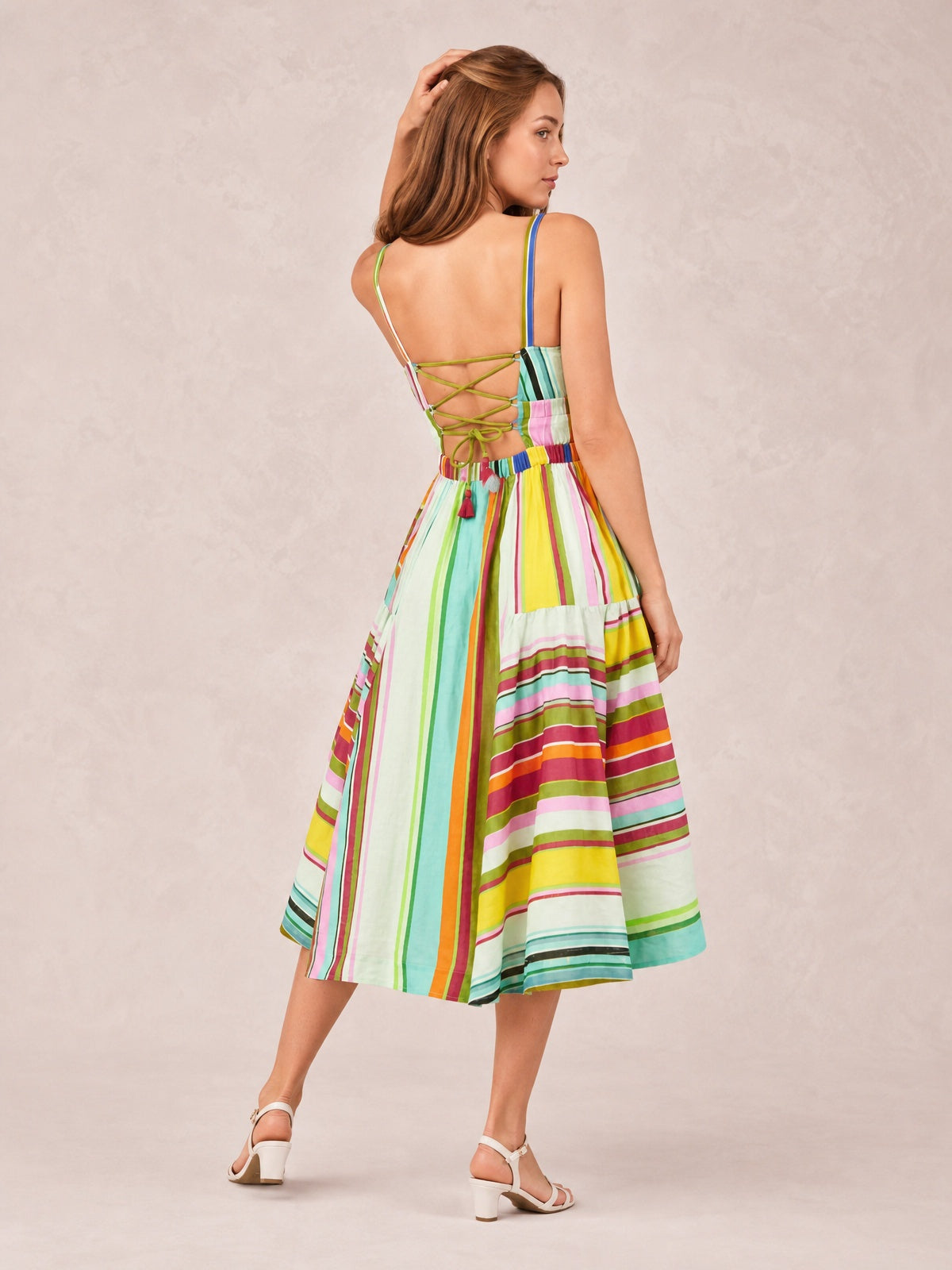 A model poses in a colorful striped spaghetti strap midi dress with an open lace-up back, defined waistband, and flowing flared skirt.