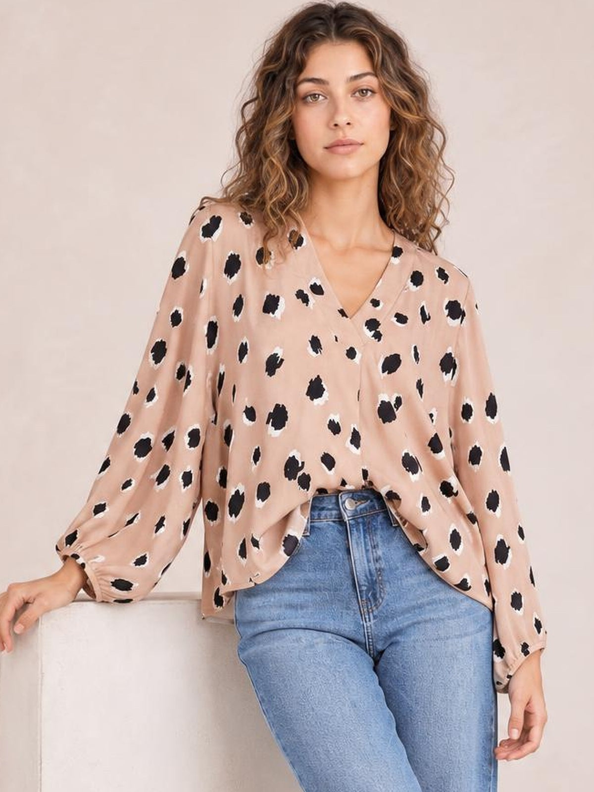 Model leans against a seat wearing a blush pink animal print blouse with a V-neck, long sleeves, and a relaxed draped silhouette.