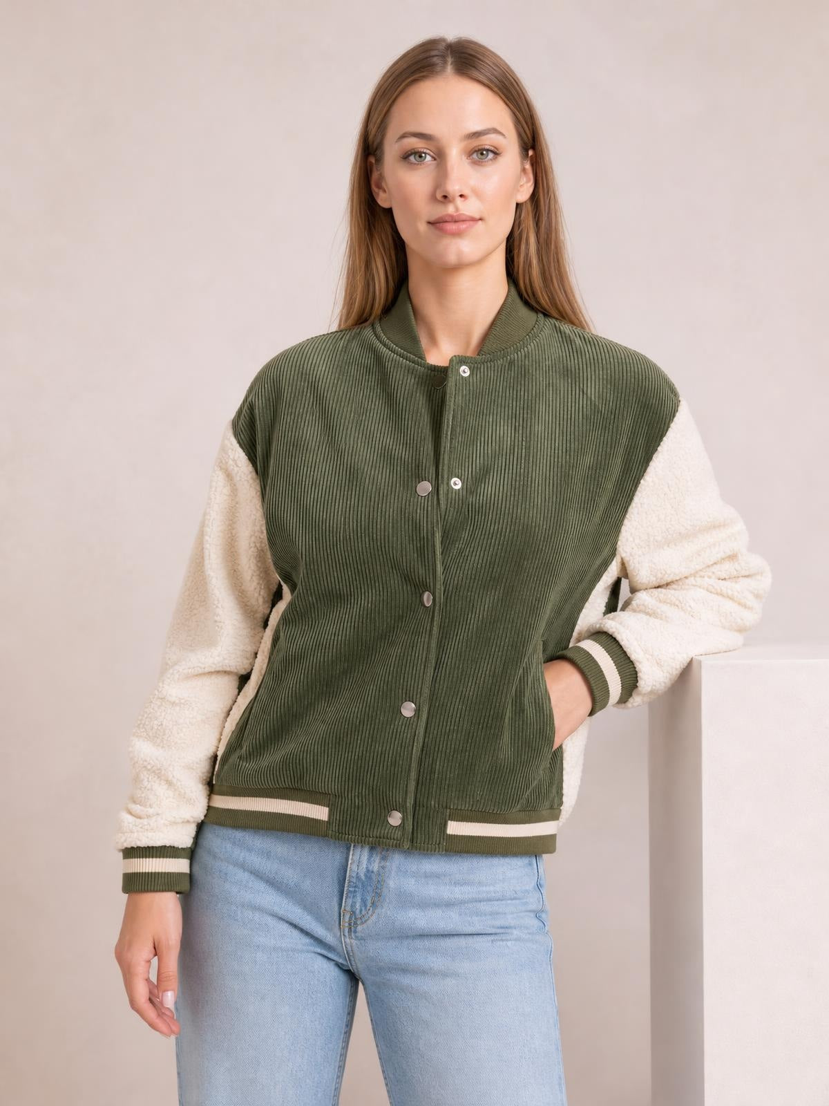 A woman models a green ribbed contrast baseball jacket with cream textured long sleeves, snap-down front, striped ribbed cuffs and hem, and a hand in a pocket.
