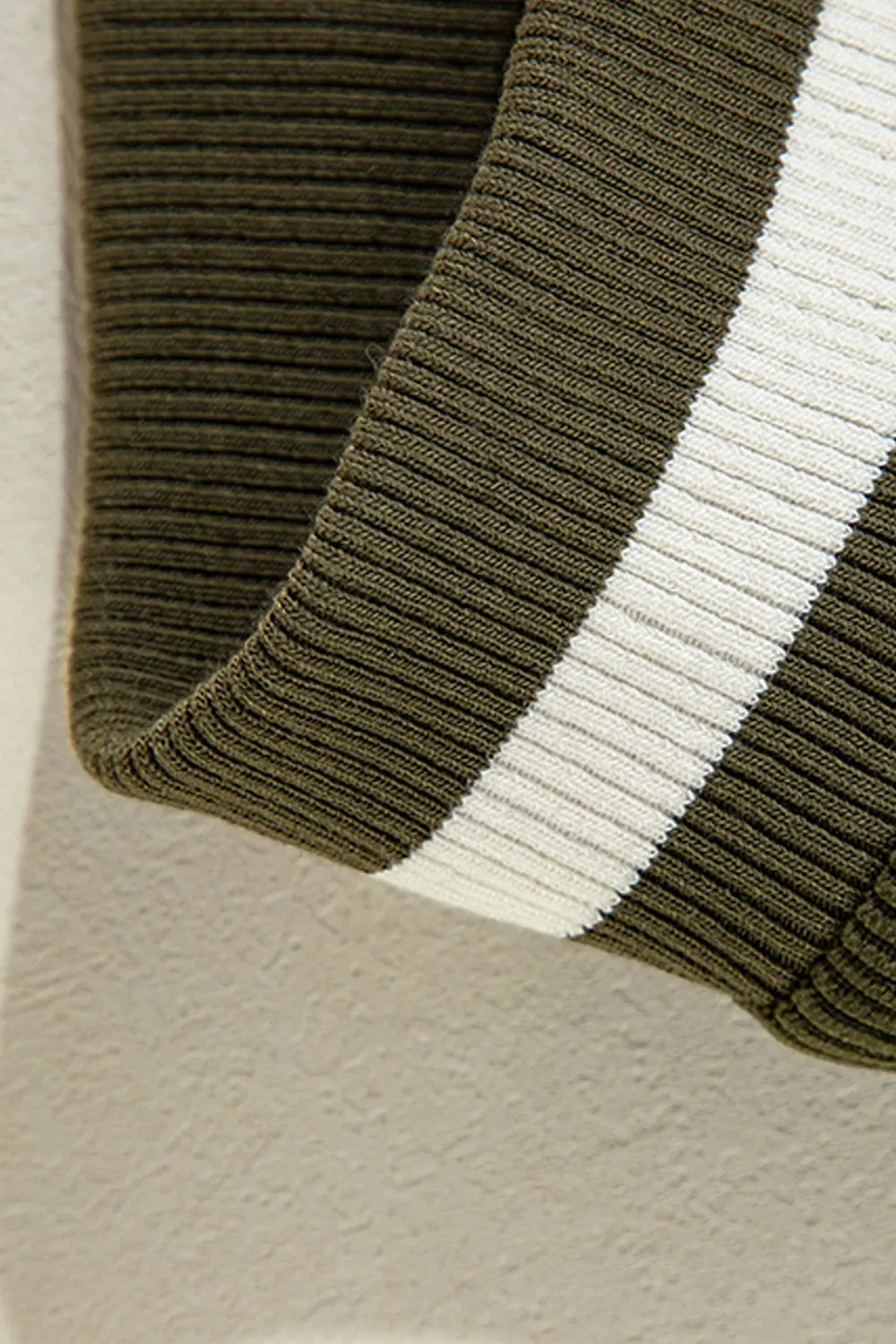 Close-up detail of an olive-green jacket with a contrasting white stripe and dark panel, showcasing ribbed trim and a curved finished edge.